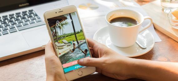 Pessoa abrindo o site do Airbnb no celular com um computador ao fundo e uma xicara de café ao lado