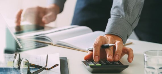 homem consultando dados em computador e realizando cálculos em calculadora